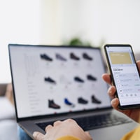 A person with a phone looking at shoes on a laptop.